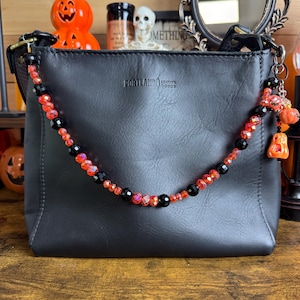 May include: A black leather shoulder bag featuring a decorative beaded strap attachment. The strap consists of alternating faceted orange and smooth black beads, with small orange pumpkin-shaped charms hanging from one side. The bag is embossed with the text PORTLAND LEATHER GOODS.