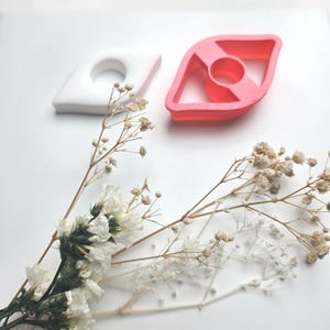 Peut inclure: Deux emporte-pièces en forme d'œil, un blanc et un rose, sont posés sur une surface blanche. Des fleurs séchées sont disposées au premier plan. Les emporte-pièces ont un design géométrique avec un centre circulaire.