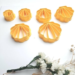 Puede incluir: Un conjunto de seis cortadores de galletas amarillos con forma de concha de diferentes tamaños. Los cortadores son de plástico y tienen un borde festoneado. Flores blancas y vegetación en el fondo.