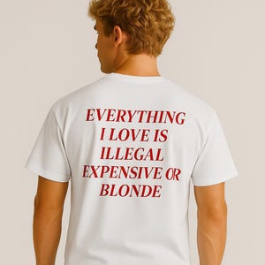 May include: White t-shirt with the red text "EVERYTHING I LOVE IS ILLEGAL EXPENSIVE OR BLONDE." The shirt is a classic crew neck style. The person in the image is wearing blue jeans.