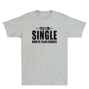 May include: A light gray t-shirt with the text "YES I'M - SINGLE - NOW IS YOUR CHANCE" printed in black. The shirt has short sleeves and a crew neck. The text is in a bold, distressed font.