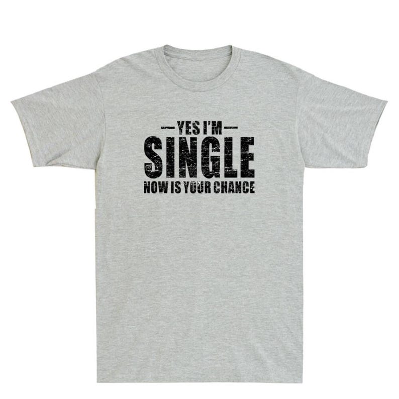 May include: A light gray t-shirt with the text "YES I'M - SINGLE - NOW IS YOUR CHANCE" printed in black. The shirt has short sleeves and a crew neck. The text is in a bold, distressed font.