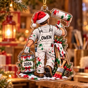 May include: A clear acrylic Christmas ornament featuring a painter wearing a Santa hat and white overalls with the name OWEN printed on the back. The figure holds a paint bucket labeled 2026 and a paint roller, surrounded by festive greenery.