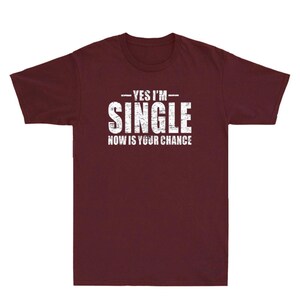 May include: A maroon t-shirt with the text "YES I'M SINGLE NOW IS YOUR CHANCE" in distressed white lettering. The shirt is a crew neck style and appears to be made of a soft material.