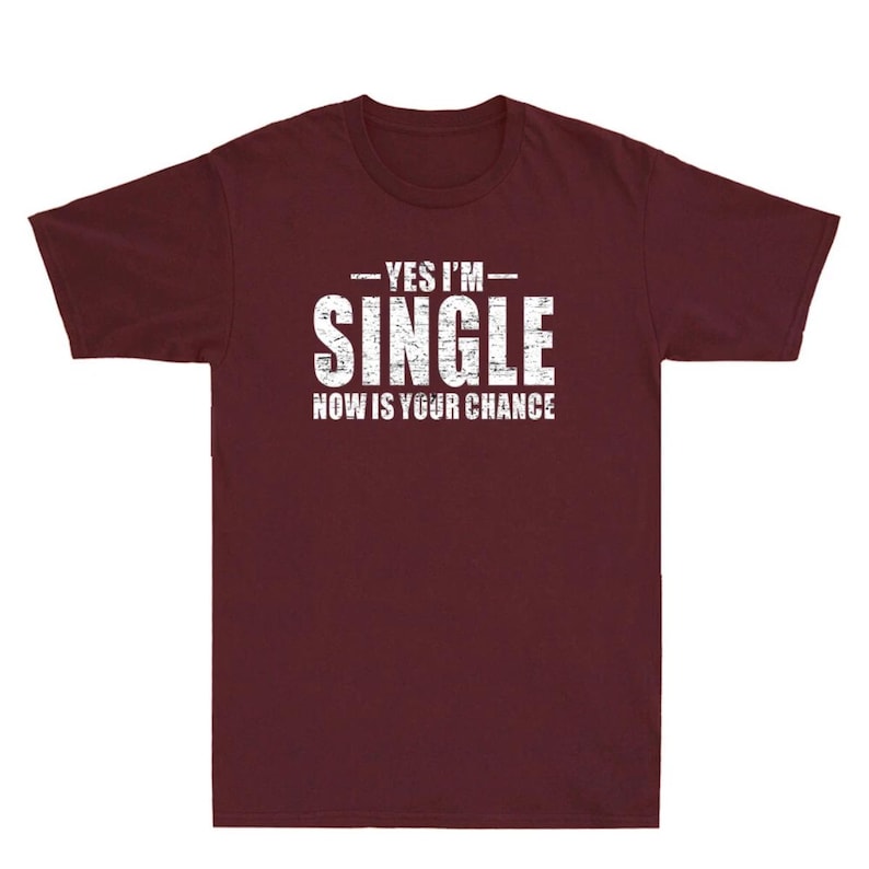 May include: A maroon t-shirt with the text "YES I'M SINGLE NOW IS YOUR CHANCE" in distressed white lettering. The shirt is a crew neck style and appears to be made of a soft material.