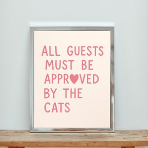 Könnte beinhalten: Ein gerahmtes Schild mit dem Text "ALL GUESTS MUST BE APPROVE[heart]D BY THE CATS" in rosa Schrift auf einem hellrosa Hintergrund.