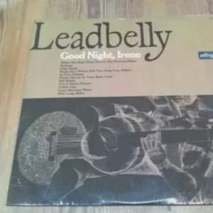 May include: A vintage vinyl record album titled "Leadbelly Good Night, Irene." The album cover features a black and white illustration of a musician playing a stringed instrument. The album is on a wooden surface.