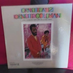 Może przedstawiać: Okładka albumu winylowego z napisem "Ornette at 12 Ornette Coleman" w kolorowych blokowych literach. Okładka albumu zawiera zdjęcie dwóch osób, z których jedna ma na sobie pomarańczowy garnitur, a druga pomarańczową kurtkę. Tytuł albumu znajduje się nad zdjęciem.