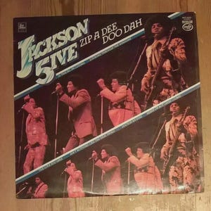 May include: Vintage vinyl record album cover for The Jackson 5ive, featuring the band members in performance. The album title is "Zip A Dee Doo Dah" with the text "14 Tracks Including" and other song titles.