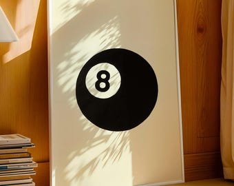 May include: A framed print of a black eight ball on a cream background. The artwork is in a white frame and leans against a wooden wall. A lamp and a stack of books are visible, suggesting a living room setting. The room has a warm, inviting atmosphere.