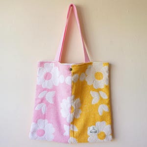 Może przedstawiać: Różowa i żółta torba typu tote z białym kwiatowym wzorem stokrotki. Torba ma różowy pasek i jest wykonana z miękkiego, teksturowanego materiału. Torba jest podzielona na dwie sekcje, różową po lewej i żółtą po prawej.