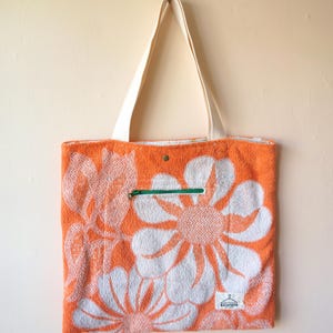 Może przedstawiać: Pomarańczowa torba typu tote z białym kwiatowym wzorem. Posiada białą rączkę i zielony zamek błyskawiczny. Torba jest wykonana z teksturowanego materiału, prawdopodobnie frotte. Mała metka jest widoczna w prawym dolnym rogu.