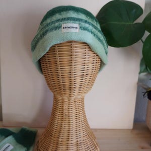 Può includere: Un berretto a righe verdi e blu con una piccola etichetta bianca con la scritta "RE-WIRED". Il cappello è esposto su una testa di manichino in vimini. Un paio di guanti abbinati è visibile nell'angolo in basso a sinistra.