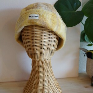 Könnte beinhalten: Ein gelb-beiger Beanie-Hut mit einem rechteckigen Etikett mit der Aufschrift "RE-WIRED". Der Hut wird auf einem Weidenkopf ausgestellt. Der Hut hat ein strukturiertes Aussehen und einen umgeschlagenen Rand. Der Hintergrund ist eine hellrosa Wand. Der Hut hat einen Durchmesser von ca. 20 cm.