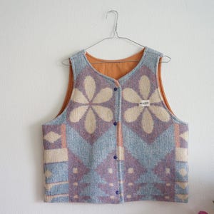 Może przedstawiać: Bezrękawnik z kwiatowym wzorem w odcieniach niebieskiego, fioletowego i kremowego. Kamizelka ma zapięcie na guziki i pomarańczową podszewkę. Wisi na metalowym wieszaku na białej ścianie.