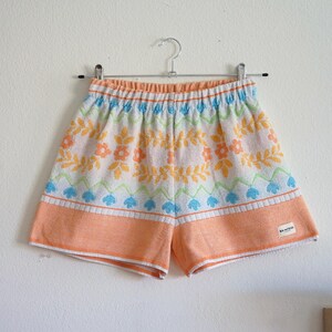 May include: Colorful, patterned shorts with an elastic waistband. The shorts feature a floral design in orange, yellow, blue, and green on a white background, with an orange border. The shorts are hanging on a metal hanger.
