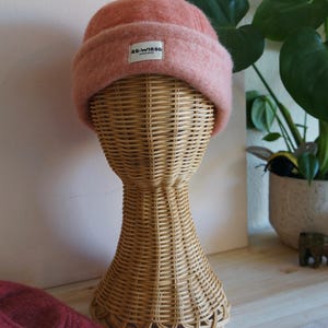 以下が含まれることがあります： 柳のマネキンの頭に展示された、折り返しのつばが付いた薄いピンク色のビーニー帽。帽子には「re-Wired」と書かれた小さな白いタグが付いています。背景には観葉植物と木製の表面が含まれています。
