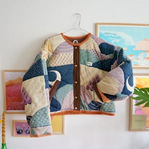 Könnte beinhalten: Eine gesteppte Jacke mit einem Patchwork-Design aus verschiedenen Stoffen in Blau-, Creme- und Lavendeltönen. Die Jacke hat einen runden Ausschnitt, eine braune Knopfleiste mit vier weißen Knöpfen sowie braune Kanten an Saum und Ärmeln.