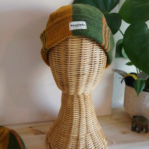 May include: A two-toned beanie hat, half mustard yellow and half green with orange stripes, is displayed on a wicker head form. The hat has a small white tag that reads "RE-WIRED". The background includes a wooden surface and a potted plant.