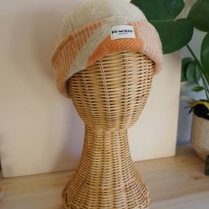 May include: A beige and orange beanie hat with a folded brim, displayed on a wicker head form. The hat features a rectangular label with the text "RE-WIRED". The hat's design includes orange and beige stripes.