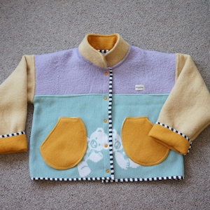 Könnte beinhalten: Bunte Kinderjacke mit einem oberen Teil in Lavendel, einem türkisfarbenen Mittelteil und beigefarbenen Ärmeln. Die Jacke hat zwei senfgelbe Taschen und einen Kragen. Die Manschetten und der Saum sind mit schwarz-weißen Streifen versehen.