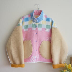 Op de afbeelding: Kleurrijk fleecejack met pastelkleuren en een uniek design. De jas heeft een roze body, beige mouwen en een geruit patroon op de schouders. De kraag en manchetten zijn geel, met twee afgeronde zakken.
