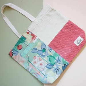 Może przedstawiać: Torba typu tote z patchworku z białym uchwytem. Torba posiada sekcje z kwiatowym, czerwono-białą kratą i białym materiałem. Mała etykieta z logo wieszaka jest wszyta na czerwono-białej kratce.