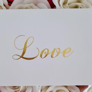 May include: A white card with the word "Love" in gold script. The card is surrounded by a background of red and white roses, creating a romantic and elegant aesthetic. Ideal for weddings or Valentine's Day.