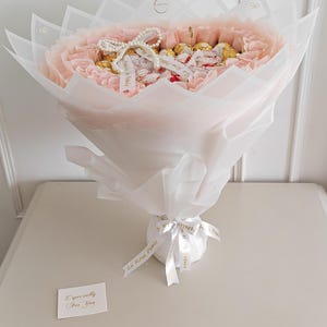 May include: A bouquet of pink flowers and chocolates wrapped in white and pink paper. The bouquet is tied with a white ribbon that has gold text. A small card that says "Especially For You" is included.