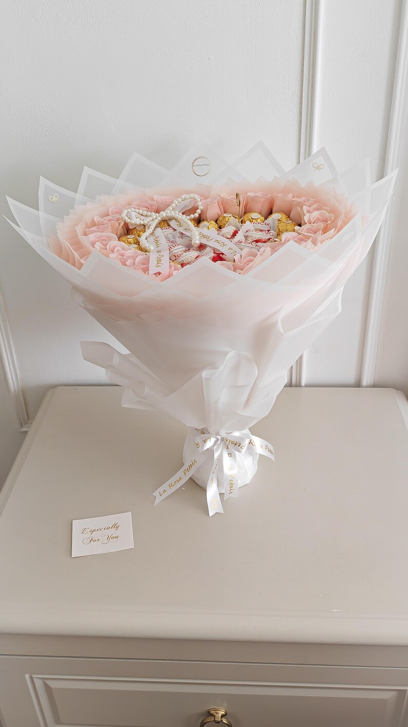 May include: A bouquet of pink flowers and chocolates wrapped in white and pink paper. The bouquet is tied with a white ribbon that has gold text. A small card that says "Especially For You" is included.