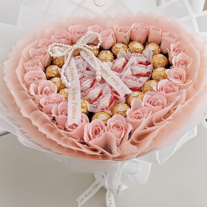 May include: A bouquet of pink roses and gold-wrapped chocolates, wrapped in layers of pink and white paper. The bouquet is tied with a white ribbon and a pearl necklace. A small card reads "Especially For You".