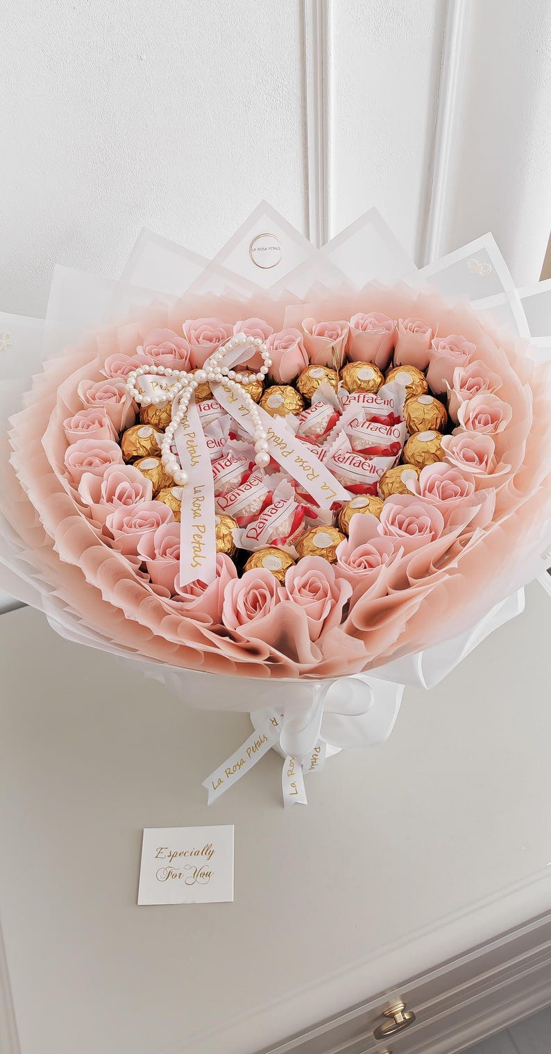 May include: A bouquet of pink roses and gold-wrapped chocolates, wrapped in layers of pink and white paper. The bouquet is tied with a white ribbon and a pearl necklace. A small card reads "Especially For You".