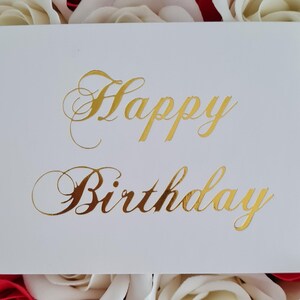 May include: A white greeting card with gold foil lettering that reads "Happy Birthday." The card is surrounded by red and white roses. The script font adds a touch of elegance to the birthday message.