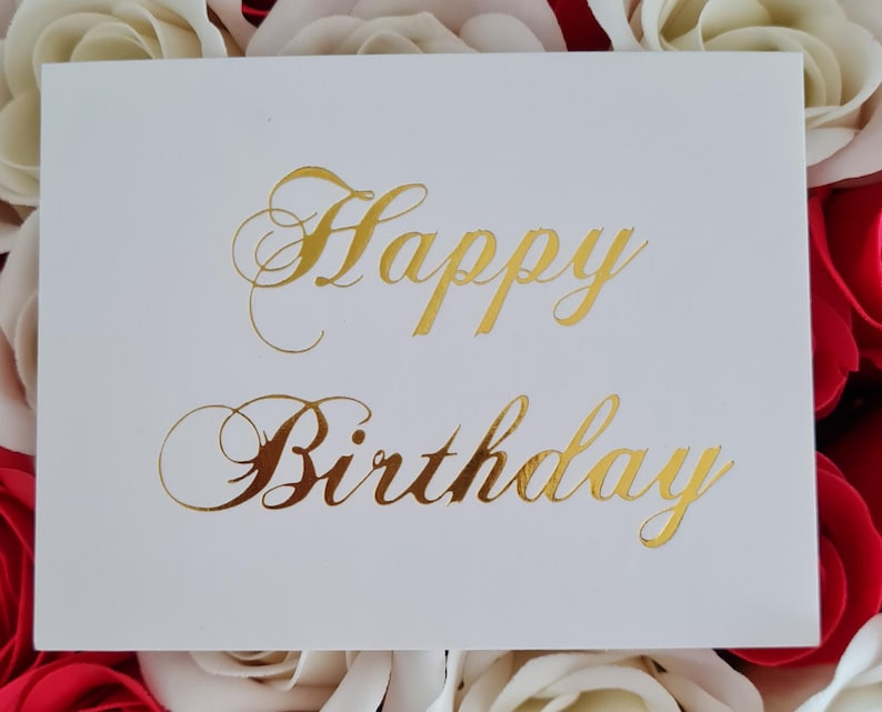 May include: A white greeting card with gold foil lettering that reads "Happy Birthday." The card is surrounded by red and white roses. The script font adds a touch of elegance to the birthday message.