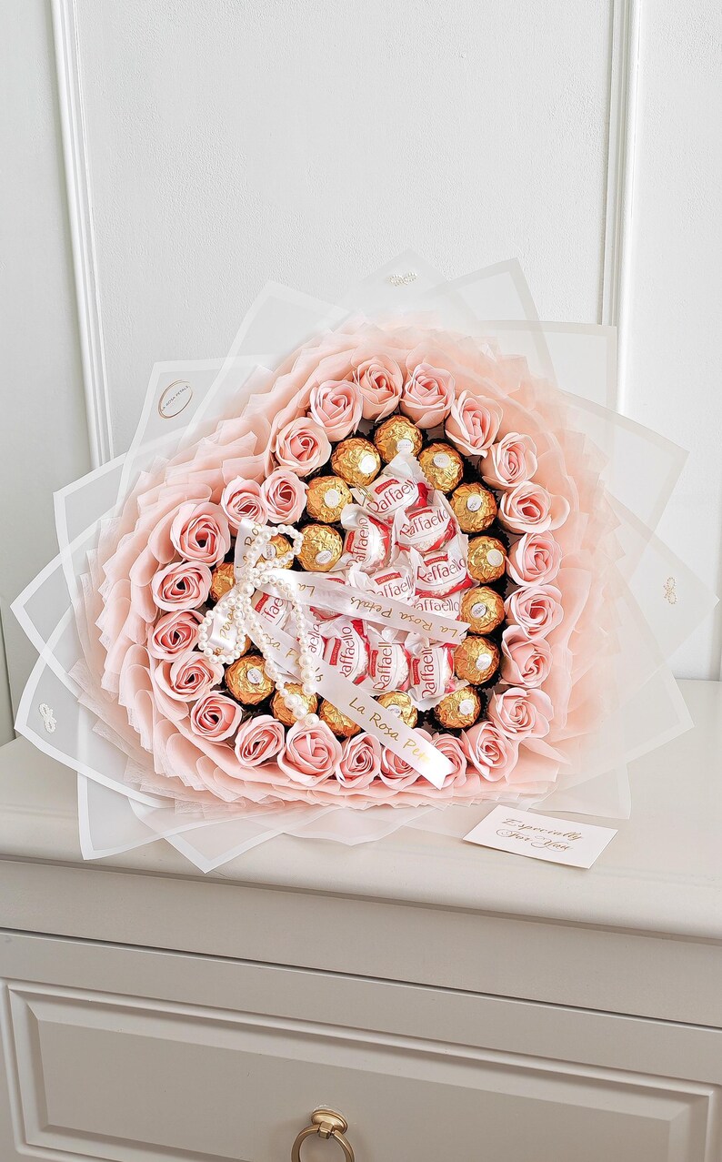 May include: A heart-shaped bouquet of pink roses and gold-wrapped chocolates. The bouquet is wrapped in white and pink paper, tied with a ribbon that reads "La Rosa Petals". A small card is included.