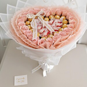 May include: A bouquet of light pink roses and gold-wrapped chocolates, arranged in a heart shape and wrapped in white and pink paper. The bouquet is tied with a white ribbon and a pearl necklace. A small card reads "Especially For You."