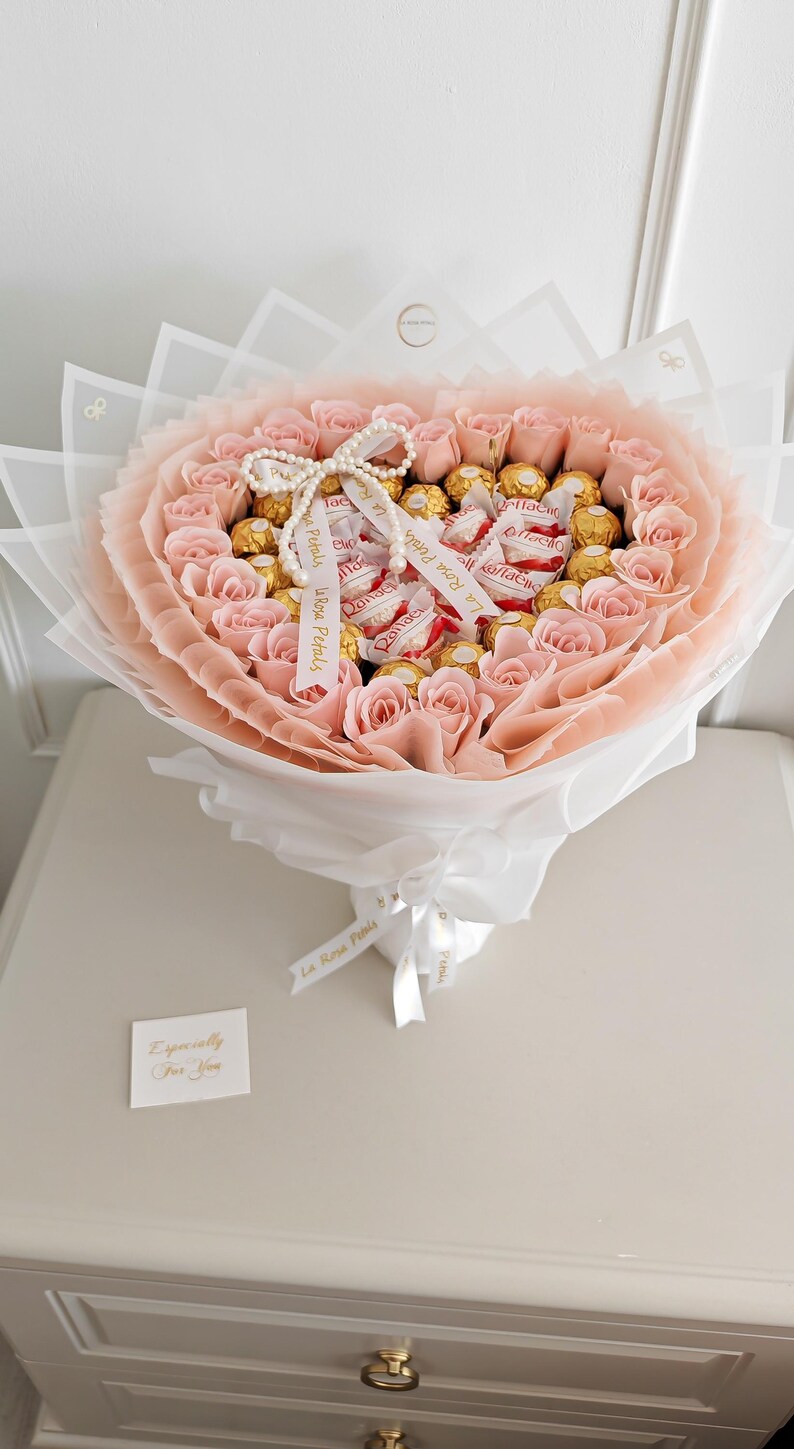 May include: A bouquet of light pink roses and gold-wrapped chocolates, arranged in a heart shape and wrapped in white and pink paper. The bouquet is tied with a white ribbon and a pearl necklace. A small card reads "Especially For You."