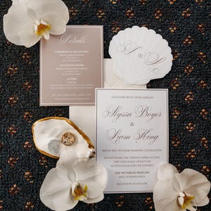 May include: A wedding invitation suite featuring white cards with black text, a shell-shaped card with the initials A & L, a gold-rimmed oyster shell holding two gold rings, and three white orchid flowers arranged on a dark blue patterned surface.