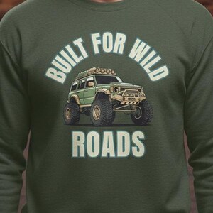 May include: A dark green sweatshirt featuring the text "BUILT FOR WILD ROADS" and an illustration of a green off-road vehicle. The background shows a desert landscape with cacti and a sunset.