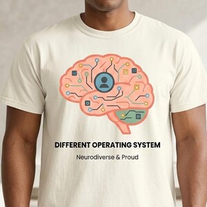 May include: Cream-colored t-shirt featuring a graphic of a brain with circuit-like lines and a person icon. The text reads "DIFFERENT OPERATING SYSTEM" and "Neurodiverse & Proud" in black font. The shirt is a crew neck style.