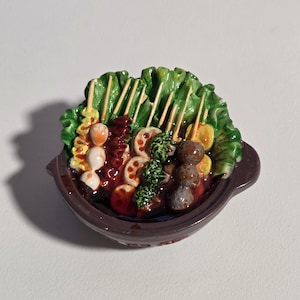 Può includere: Un piatto in ceramica con un assortimento colorato di cibi, tra cui spiedini, verdure e vari ingredienti. Il piatto marrone ha una guarnizione di foglie verdi e gli alimenti sono dettagliati con colori vivaci.