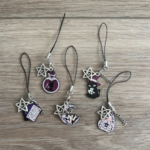 May include: A collection of six charm pendants with black cords and silver-tone metal accents. The charms feature various designs, including a potion bottle, a black cat in a witch hat, a crescent moon, and a sugar skull. The charms are set against a wooden background.
