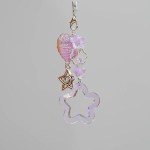 Puede incluir: Un delicado dije con forma de flor morada transparente, una estrella plateada y una cuenta en forma de corazón con tonos iridiscentes rosas y naranjas. El dije está adornado con pequeñas cuentas moradas y un cierre plateado.