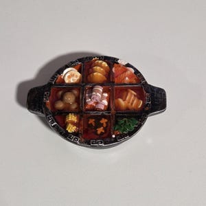Può includere: Spilla rotonda color argento con un design dettagliato. La spilla presenta un pannello centrale diviso in nove quadrati, ciascuno contenente un alimento in miniatura. Gli alimenti includono gnocchi, panini e verdure.