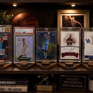 Puede incluir: Una estanter&iacute;a de madera que muestra tarjetas de baloncesto en fundas protectoras, cada una sobre un soporte de madera. Un bal&oacute;n de baloncesto y una foto enmarcada son visibles en el fondo. Libros y un letrero decorativo tambi&eacute;n est&aacute;n en el estante.