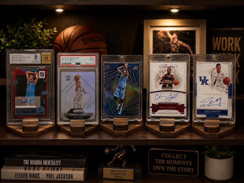 Puede incluir: Una estanter&iacute;a de madera que muestra tarjetas de baloncesto en fundas protectoras, cada una sobre un soporte de madera. Un bal&oacute;n de baloncesto y una foto enmarcada son visibles en el fondo. Libros y un letrero decorativo tambi&eacute;n est&aacute;n en el estante.