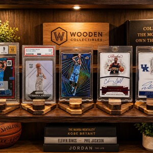 Puede incluir: Una exhibici&oacute;n de cromos de baloncesto en fundas protectoras, cada uno en un soporte de madera. Las tarjetas presentan aut&oacute;grafos de jugadores y est&aacute;n dispuestas en una estanter&iacute;a con una pelota de baloncesto y libros. La estanter&iacute;a tambi&eacute;n muestra el texto "Wooden Collectibles".