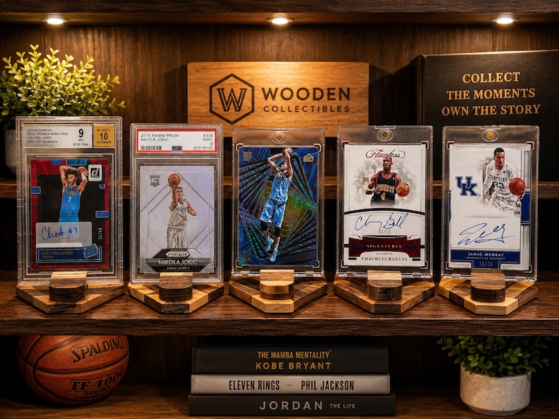 Puede incluir: Una exhibici&oacute;n de cromos de baloncesto en fundas protectoras, cada uno en un soporte de madera. Las tarjetas presentan aut&oacute;grafos de jugadores y est&aacute;n dispuestas en una estanter&iacute;a con una pelota de baloncesto y libros. La estanter&iacute;a tambi&eacute;n muestra el texto "Wooden Collectibles".