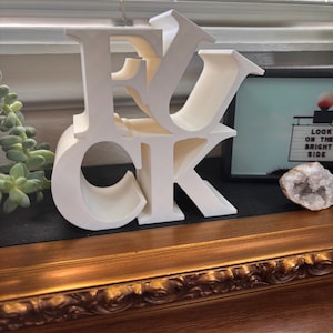 Puede incluir: Un objeto decorativo blanco impreso en 3D que presenta las letras F, U, C y K apiladas en forma de bloque. Las letras están dispuestas con la F y la U colocadas sobre la C y la K, creando una escultura tipográfica audaz y autónoma.