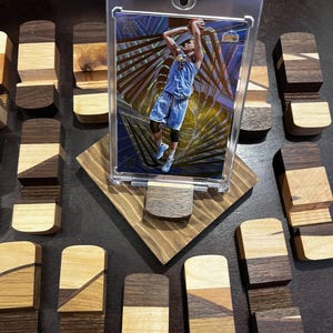 Puede incluir: Una tarjeta de baloncesto en una funda protectora transparente, exhibida en un soporte de madera. El soporte est&aacute; hecho de varios tonos de madera y est&aacute; rodeado de peque&ntilde;os bloques de madera. La tarjeta presenta a un jugador con uniforme azul.